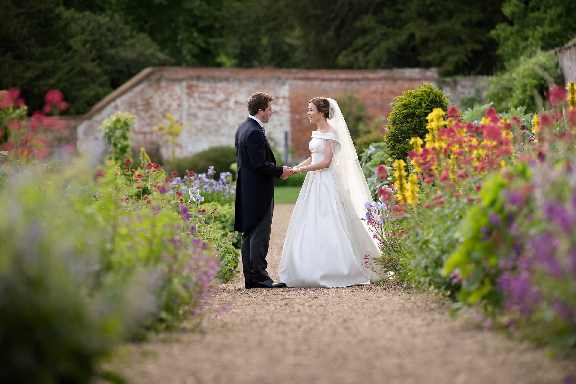 Bride and groom holding hands and chatting in a colourful flower border in the walled garden at Kelmarsh Hall