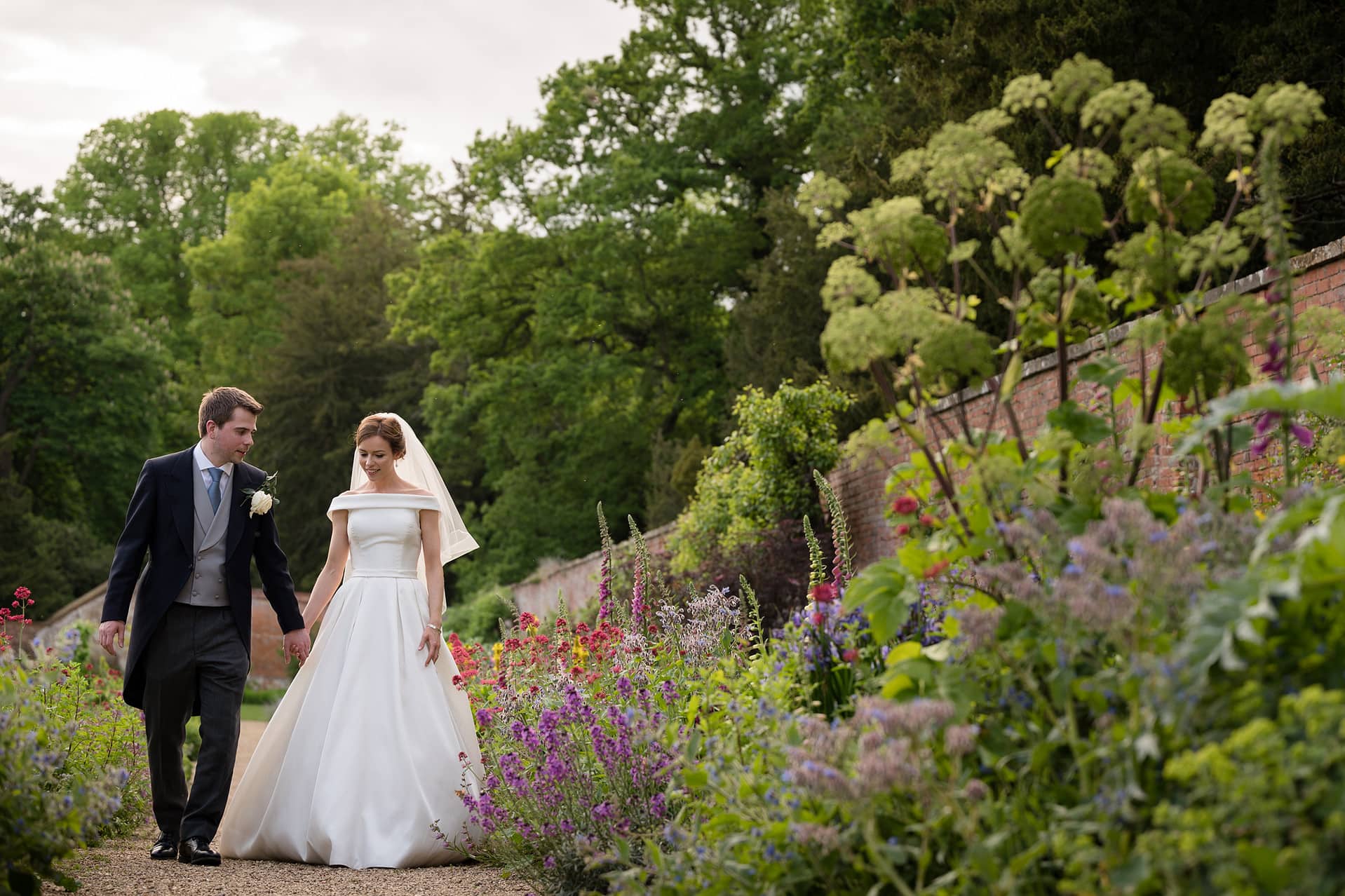 Bride and groom walking through in a colourful flower border in the walled garden at Kelmarsh Hall