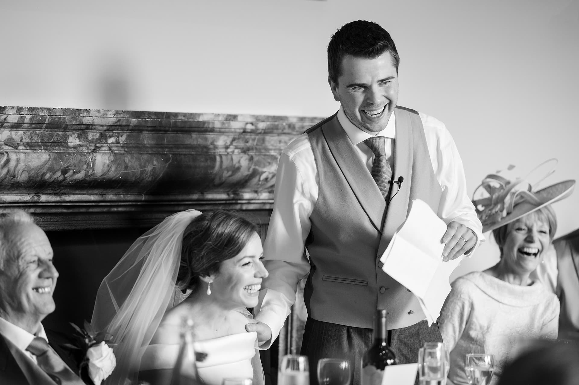 The groom laughing and reaching out to touch the bride's shoulder as he makes a mistake during his speech