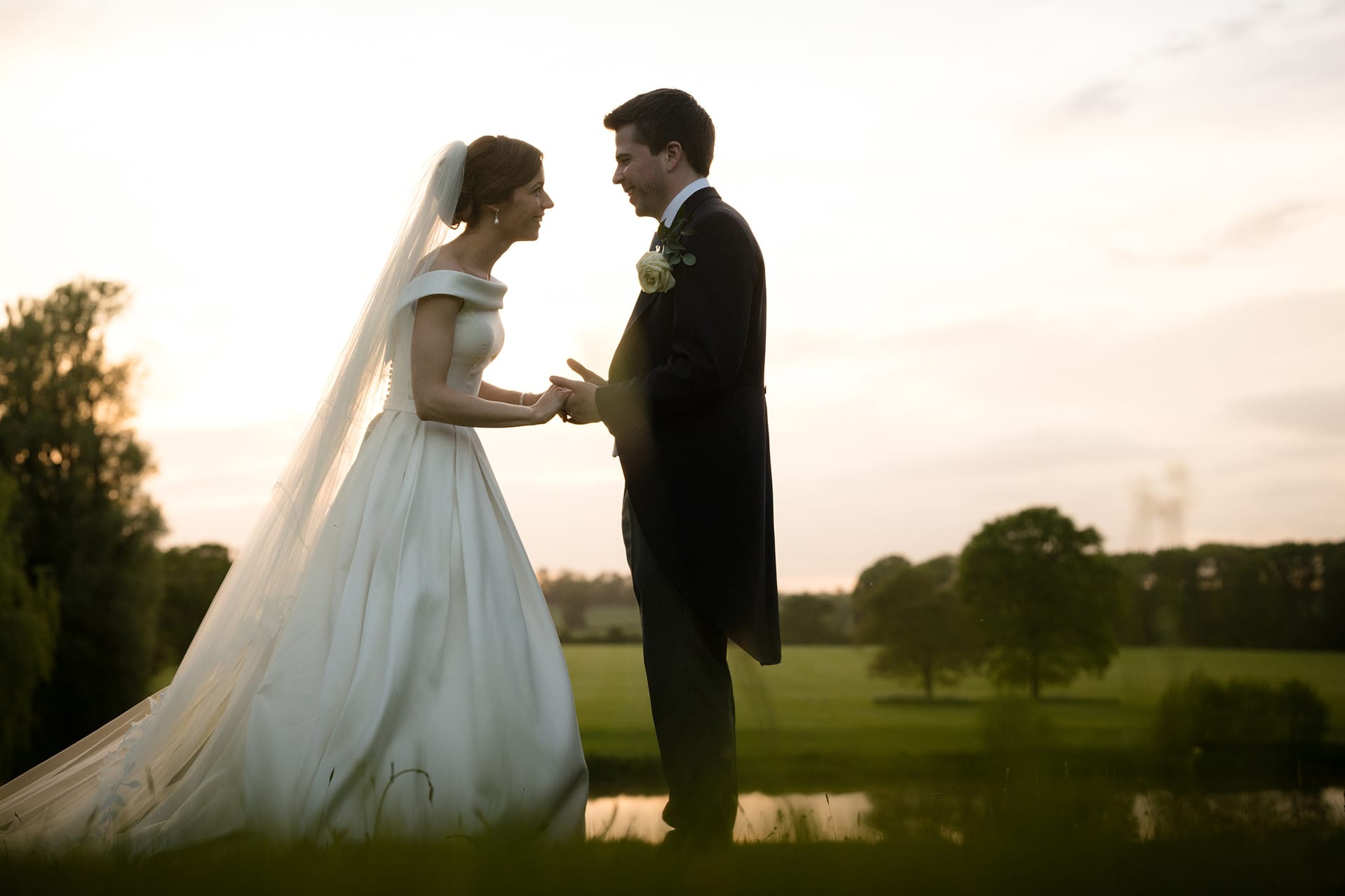 The bride and groom chatting together and holding hands during golden hour by the lake at Kelmarsh Hall