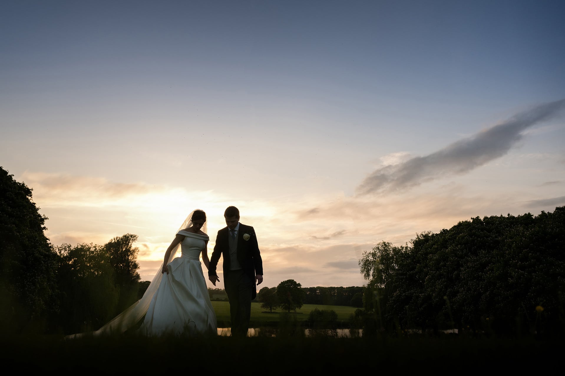 A semil-silhouette of the bride and groom walking by the lake at Kelmarsh Hall at sunset