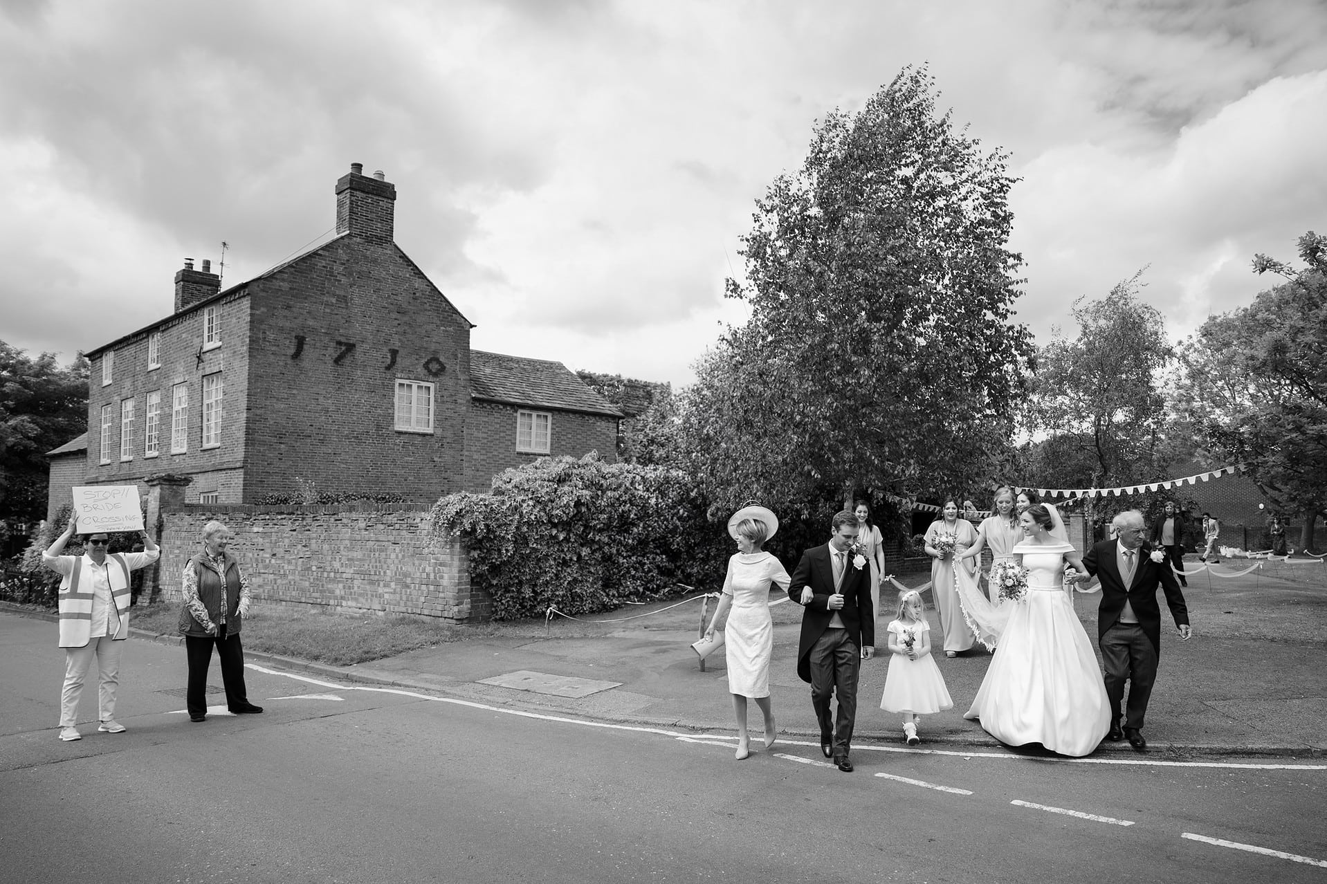 A neighbour holding up a 'Stop, Bride Crossing' sign as the bride and her entourage cross the road