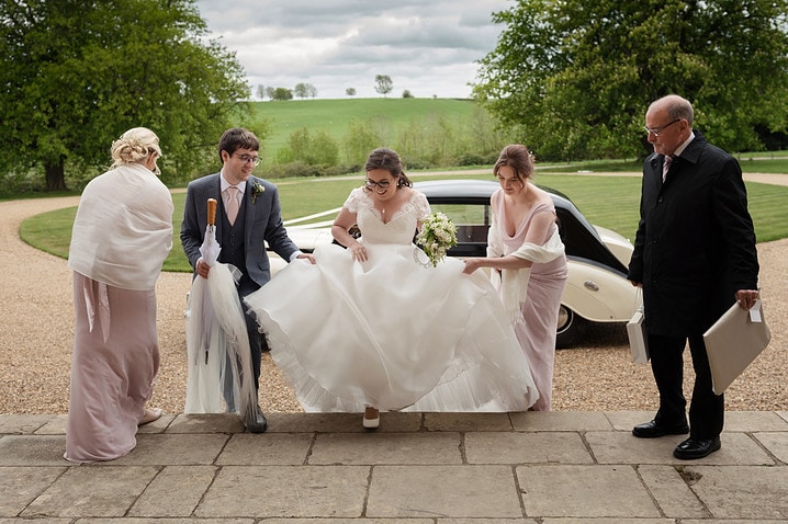 A groom and bridesmaids helping the bride with her dress as they walk up the front steps at Kelmarsh Hall