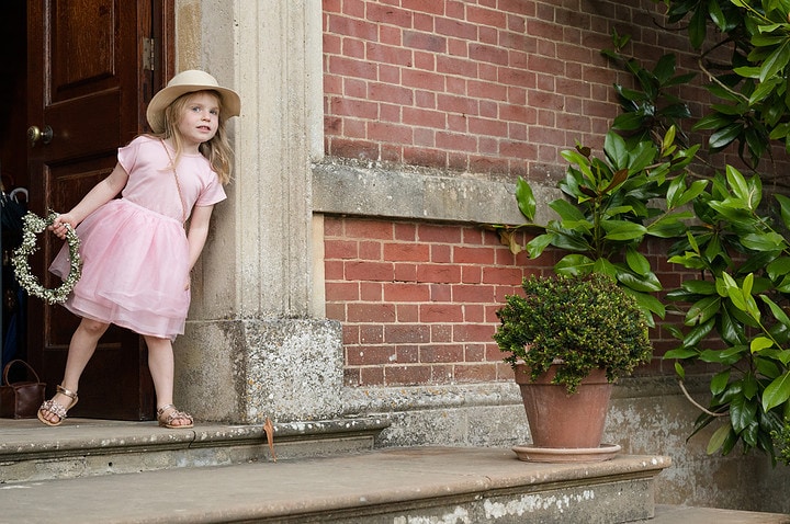 A little girl wearing a straw hat and holding a flower hoop peers out of a doorway at Kelmarsh Hall