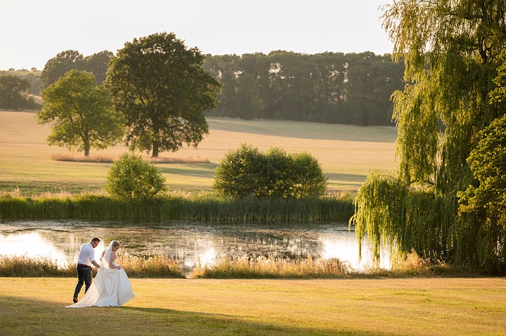 A bride and groom walking along the lakeside at Kelmarsh Hall during golden hour