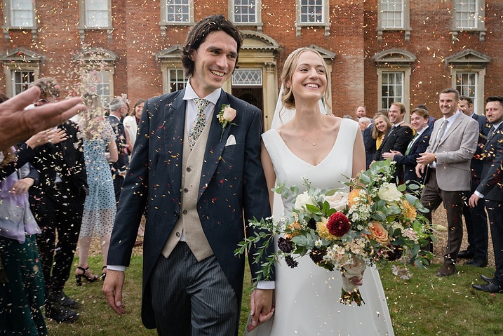 Bride and groom with big smiles as their guests throw confetti over them in front of Kelmarsh Hall
