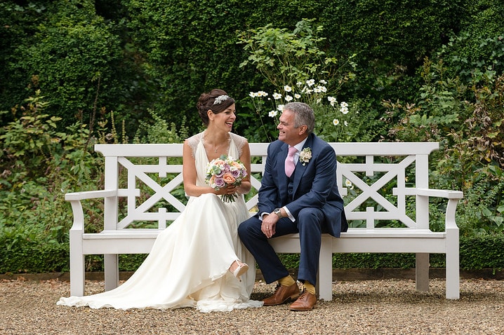 A bride and groom sitting together chatting on a white wooden bench in the sunken garden at Kelmarsh Hall