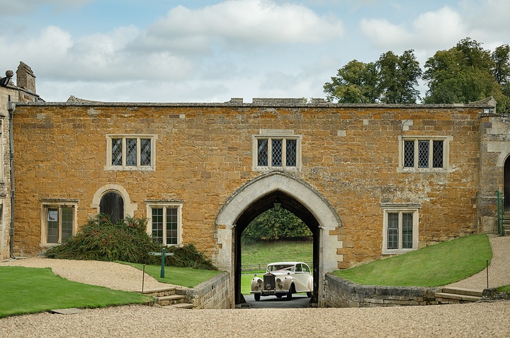 A white vintage wedding car driving through the arched entrance at Rockingham Castle
