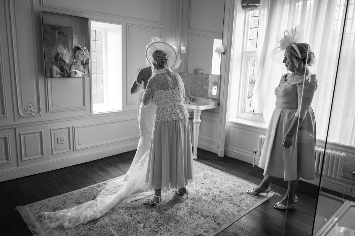 Two ladies in wedding outfits helping bride into her dress, bride reflected in mirror