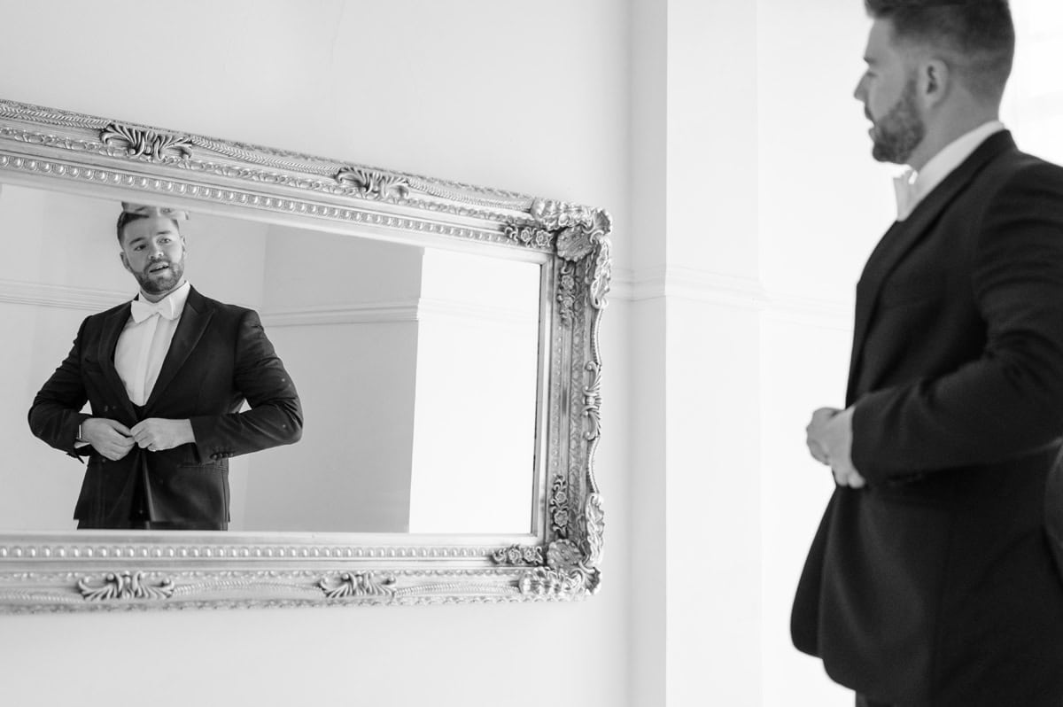 Groom doing up his suit jacket in an ornate mirror