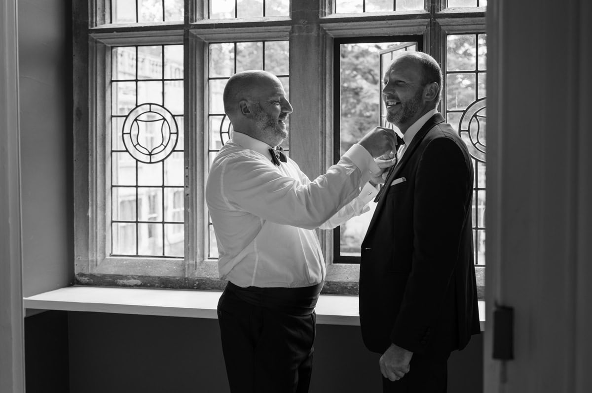 Two men helping each other with their bow ties in front of a stately window