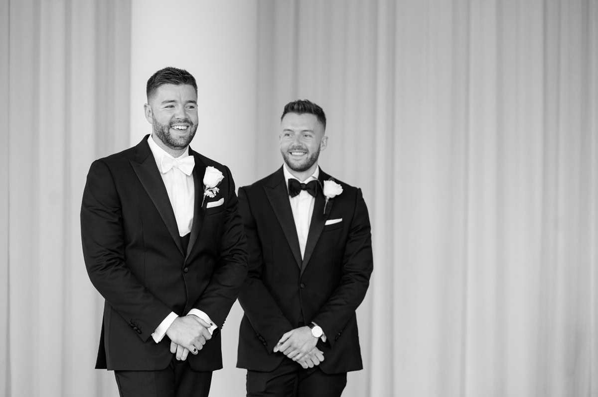 Groom and best man standing and smiling in front of a neutral draped wall as they wait for wedding ceremony to start