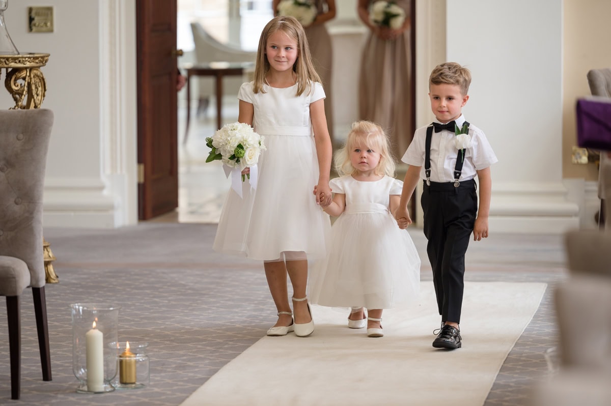 Two flower girls in white dresses and a page boy in black braces and bow tie walking down a white-carpeted aisle