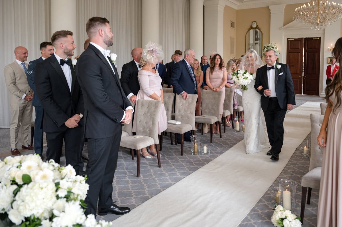 Bride walking down the aisle with her dad as the groom watches, in a grand ballroom