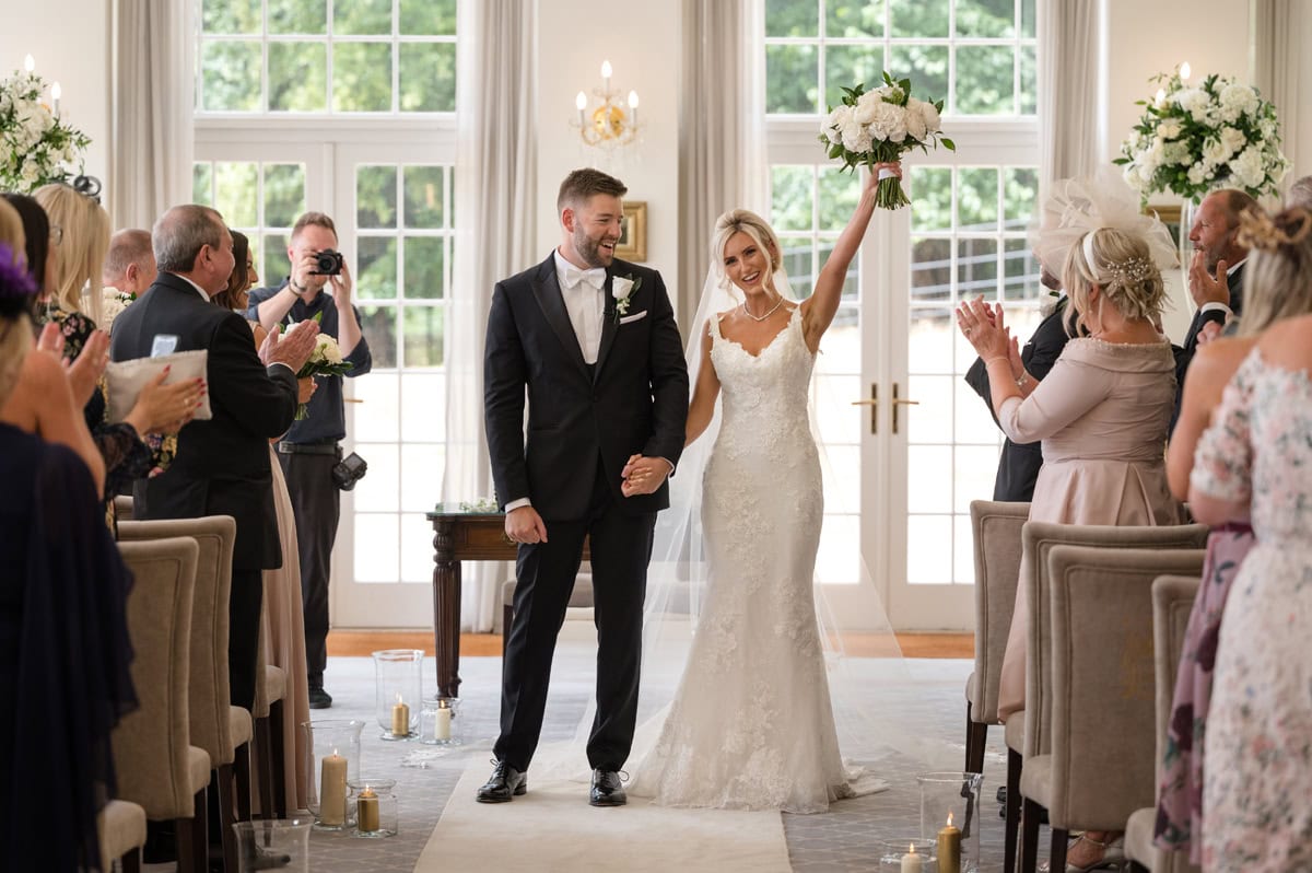 Bride triumphantly waving her bouquet, wedding guests clapping