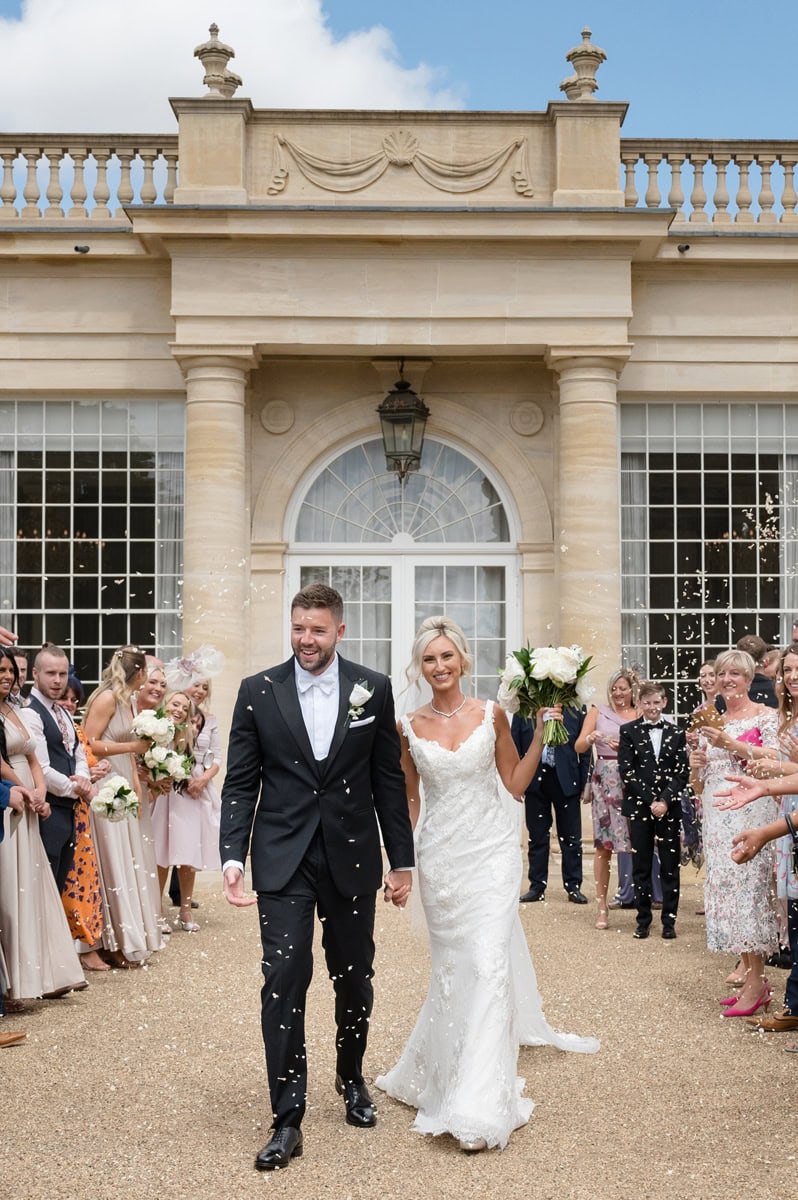 Bride and groom being covered in confetti as they walk along a path in front of a stately orangery