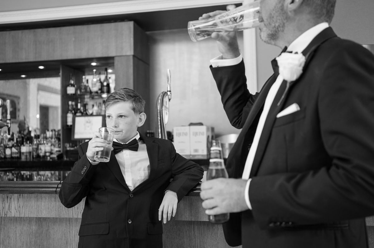 Young boy in black tie leaning against the bar as he enjoys a drink, mirroring the actions of a man doing the same with a beer next to him