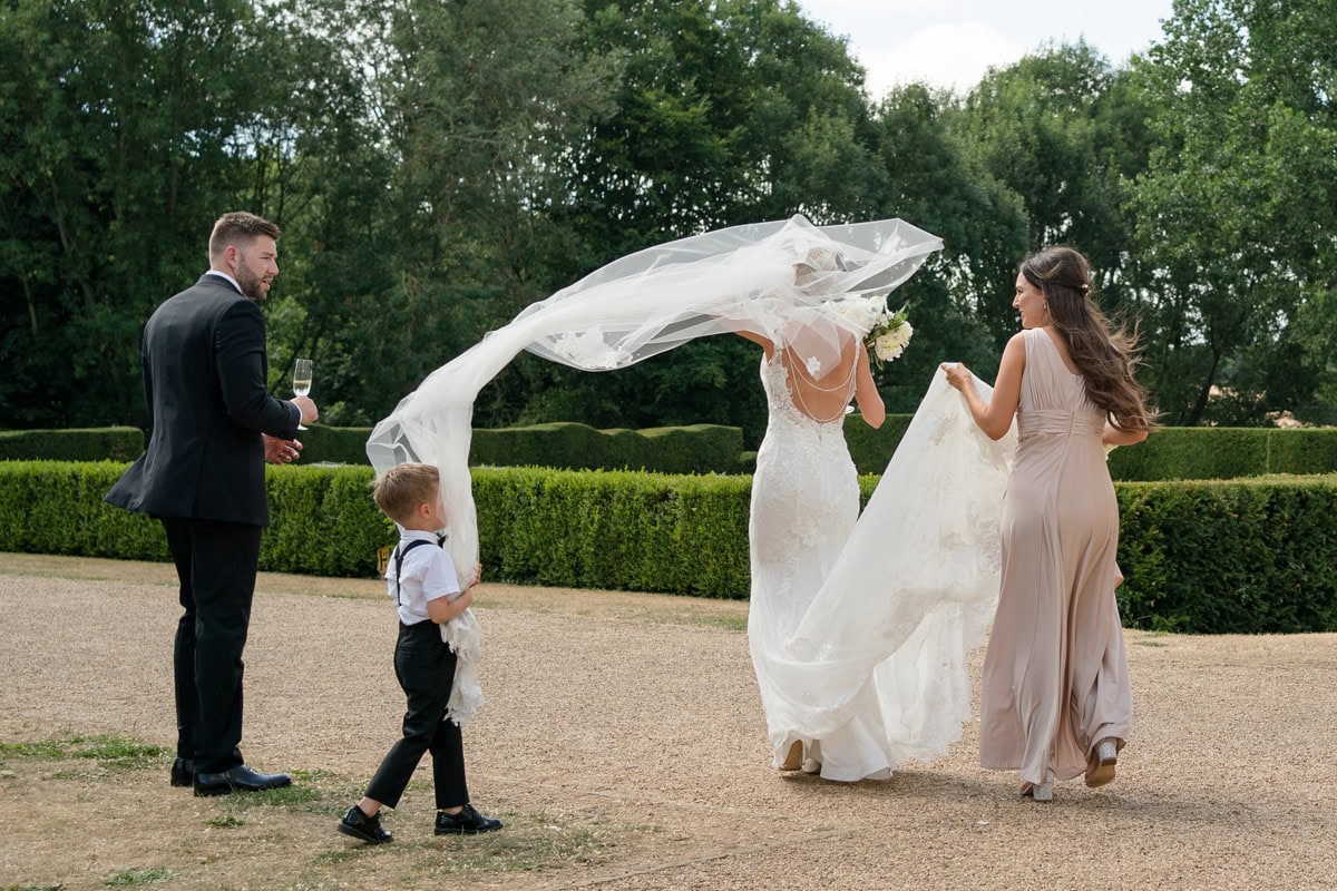 Pageboy holding onto the end of a bride's veil as it blows up in the wind into his face