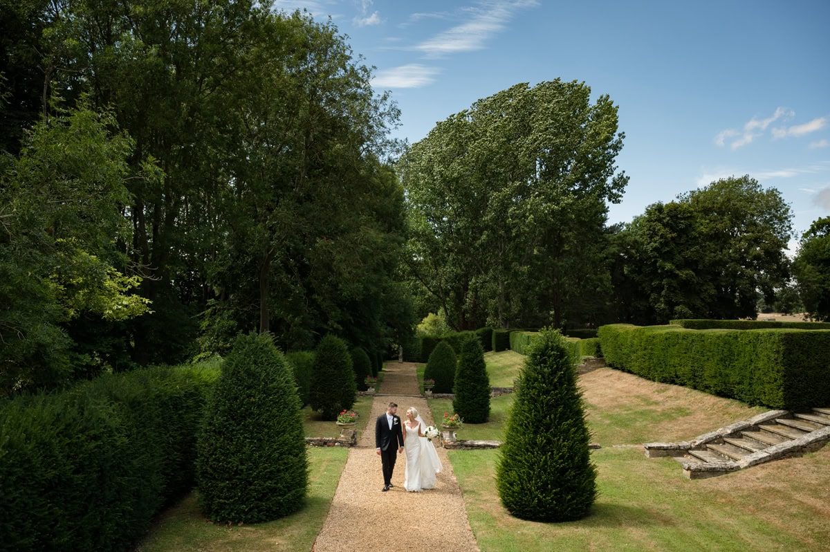 Scenic photo of a bride and groom walking down a topiary-lined path with mature trees in the background