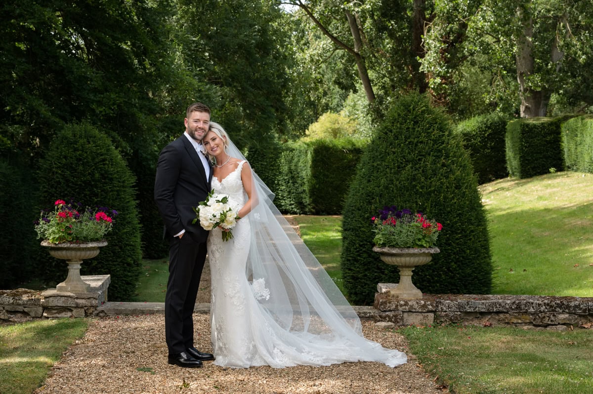Bride and groom posing for a traditional portrait in front of a topiary-lined path
