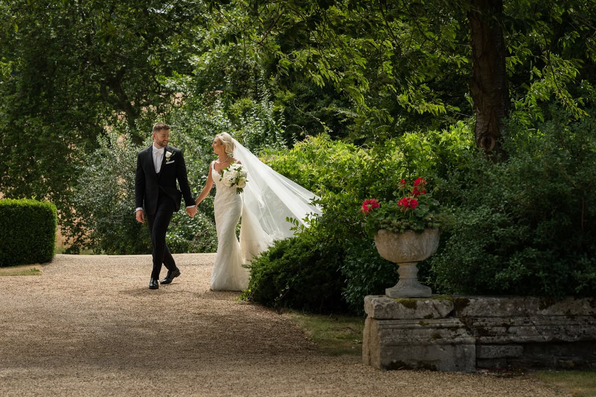 Bride and groom walking hand-in-hand around the corner of a path lined with large shrubs and trees
