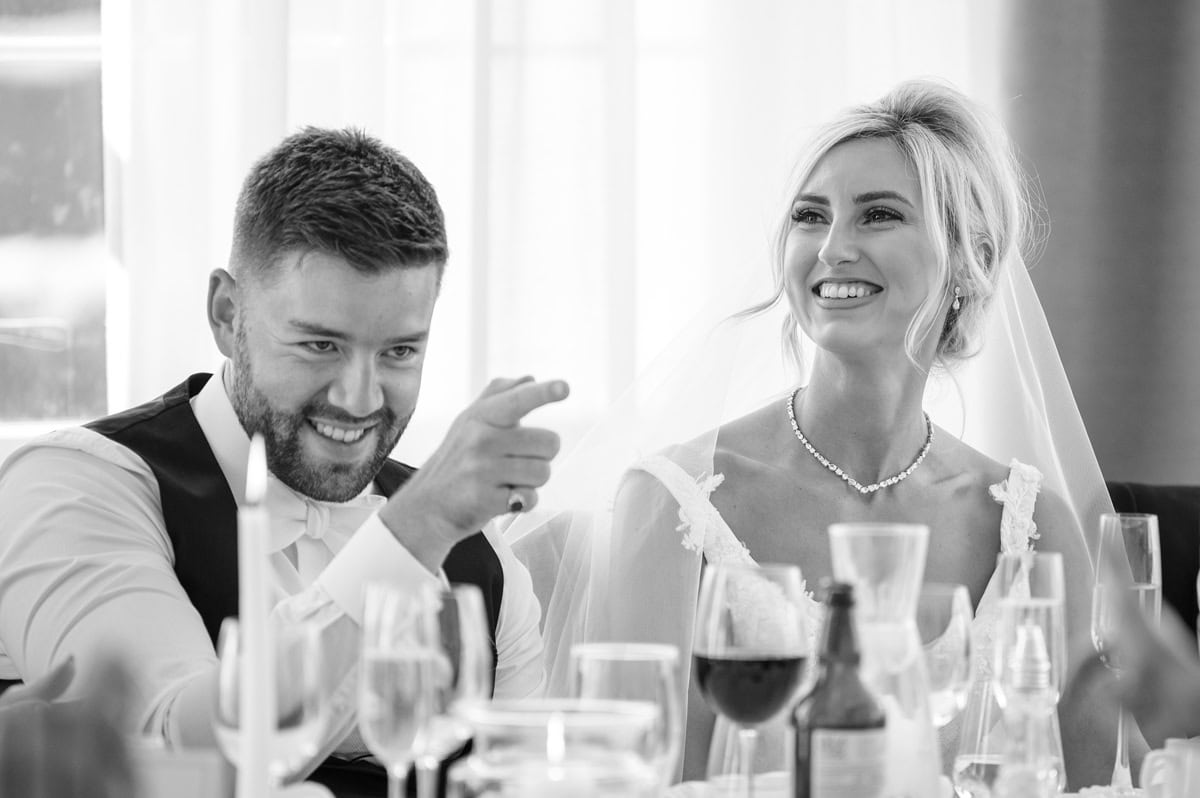 Bride smiling during wedding speech, groom pointing at someone