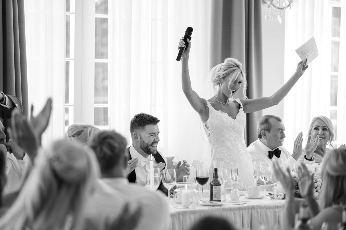 Bride giving a speech with microphone at wedding reception, guests clapping and smiling.