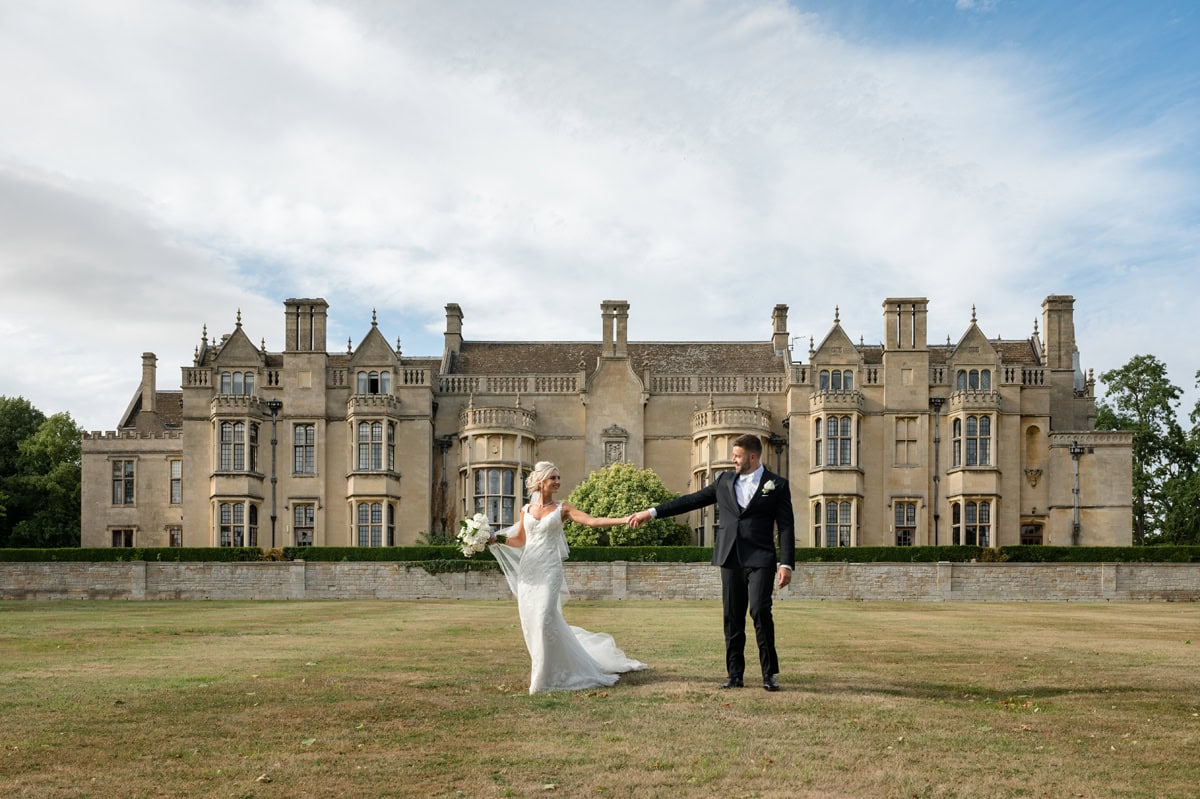 Bride and groom walking hand-in-hand with a stately Elizabethan mansion behind them
