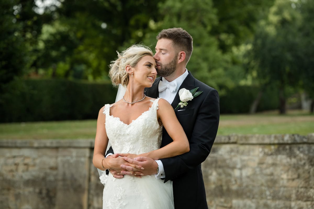 Groom gently kissing bride on the forehead in front of a stone wall and trees
