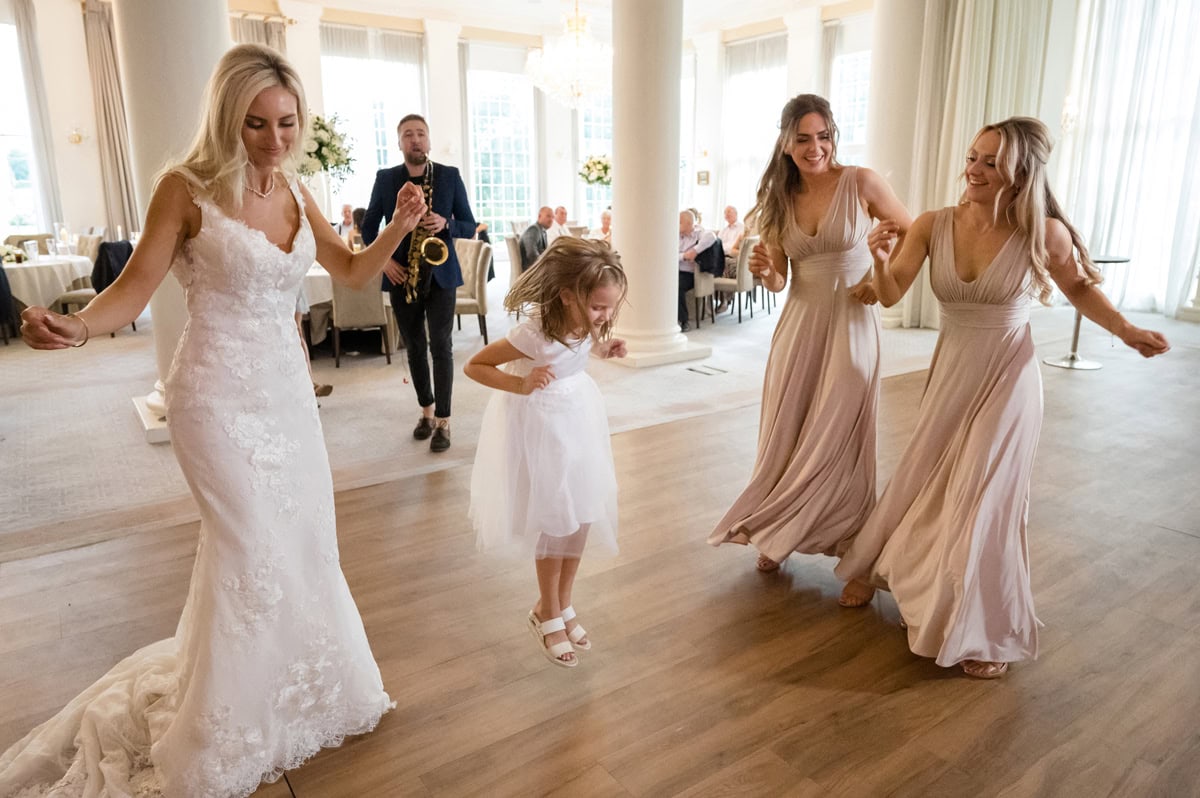 A lively scene of bride, bridesmaids, and a flower girl dancing at a wedding with a saxophone player